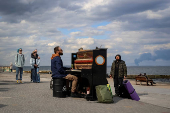 Street musician conducts an impromptu piano concert on the beach to support local residents as smoke rises over the port of Pivdennyi after Russian missiles in Odesa