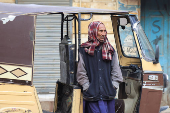 A rickshaw driver stands with his hands in a jacket while covering his head, in Karachi