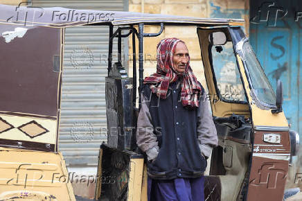 A rickshaw driver stands with his hands in a jacket while covering his head, in Karachi