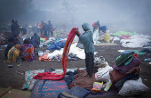 A girl folds her blanket as others sit next to a fire at an open ground on a foggy winter morning in the old quarters of Delh