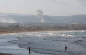 People walk at a beach as smoke rises in the background, as seen from Tyre