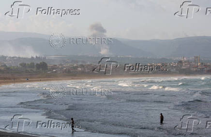 People walk at a beach as smoke rises in the background, as seen from Tyre