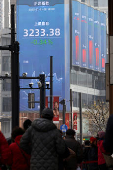 Pedestrians wait for a street signal on a sidewalk as an electronic billboard shows the Shanghai stock index in Shanghai