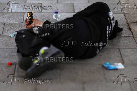 A man lies on a sidewalk with a cup for spare change near a bus stop in Tel Aviv, Israel