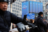 Pedestrians wait for a street signal on a sidewalk as an electronic billboard shows the Shenzhen stock index in Shanghai