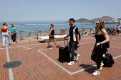 A tourist couple walk with their travel suitcases along the promenade of Las Canteras Beach in Las Palmas de Gran Canaria