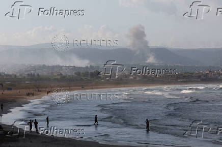 People walk at a beach as smoke rises in the background, as seen from Tyre