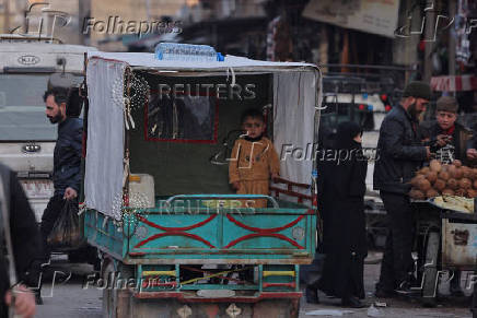 A boy stands on  the back of a vehicle, driving along the market street, in Douma