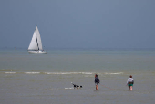 People walk through the sea with their dog in front of a sailing boat on the beach at Gravelines, France