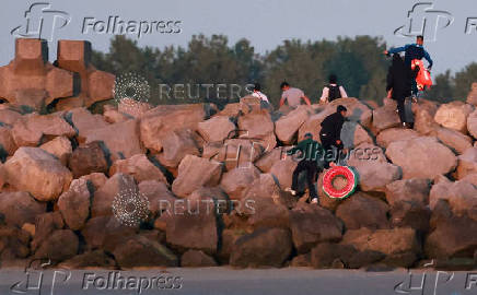 A group of migrants carrying life jackets and rubber rings climb over rocks after abandoning a dinghy whilst trying to cross the English Channel on a beach at Gravelines