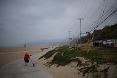 A woman walks her dogs along the beach as a truck drives down Pacific Coast Highway, in Los Angeles