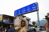 Man carries a jerrycan, on his shoulder with the signboard in the background in Chakothi Village