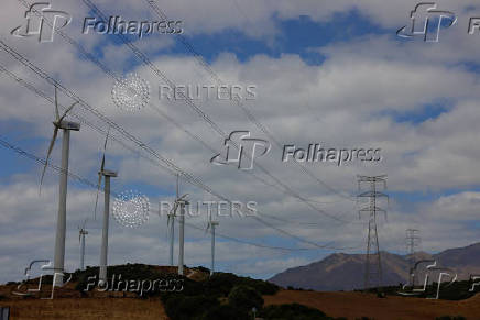 Wind turbines spin next to power lines connecting pylons of high-tension electricity, near Casares