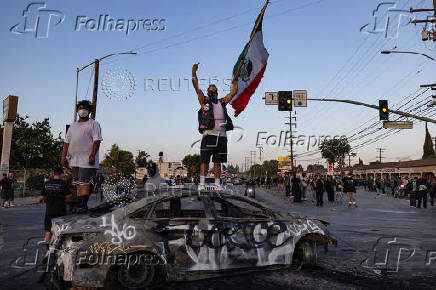 A protester holding a Mexican flag stands on a burnt car during a standoff with law enforcement in Paramount