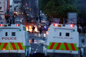 People walk near official vehicles as flames rise during a second night of riots, in Ballymena