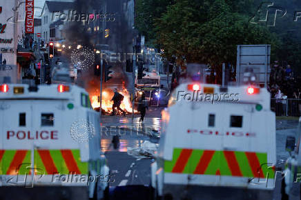 People walk near official vehicles as flames rise during a second night of riots, in Ballymena