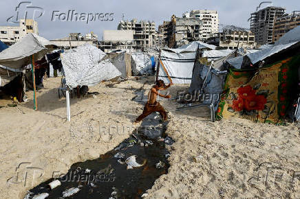 A Palestinian boy, displaced by the Israeli offensive, jumps over a puddle of wastewater on beach as damaged buildings appear in the background, amid summer heat, in Gaza City